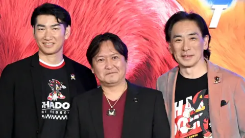 Getty Images Tiga pria Jepang berdiri bersama sambil tersenyum untuk difoto di pemutaran perdana film. Di belakangnya terdapat latar belakang yang menampilkan karakter dalam film Sonic. Ketiga pria tersebut masing-masing mengenakan pakaian yang menampilkan Shadow the Hedgehog, yang tampil dalam film tersebut. Satomi dan Utsumi memiliki Shadow di kaos serta lencana kerah, sedangkan Iizuka memiliki kalung Shadow.